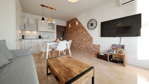 Un moderno salón con una mesa de centro de madera, un sofá gris y un área de comedor con sillas blancas. Se ve una pared de ladrillo y un televisor al fondo.