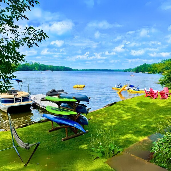 A lakeside dock with boats, a grassy area with Adirondack chairs, and a view of the lake and distant shoreline.