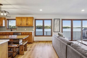 A modern kitchen with wooden cabinets, a granite countertop, and a dining area. There is a large window with a view of a lake.