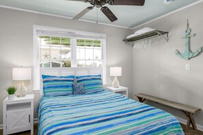 Un dormitorio con cama, dos mesitas de noche, ventilador de techo, ventana con persianas, estante montado en la pared y un banco.