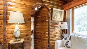 A log cabin interior with a wooden wall, a white sofa with patterned cushions, a table lamp, and a framed picture.