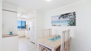 A bright kitchen with white cabinets, a wooden dining table with chairs, and a painting of a coastal scene. - Kirra Gardens 17 - Kirra Point Beachfront (Coolangatta)