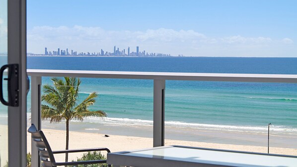 Blick auf einen Strand mit Stadtansicht in der Ferne, eine Palme und ein Stuhl auf dem Balkon.