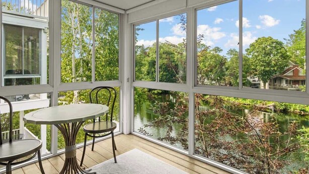 A sunlit room with large windows overlooking a lake, furnished with a table and chairs. - Midtown Riverfront Condo Extended Stays (Traverse City)