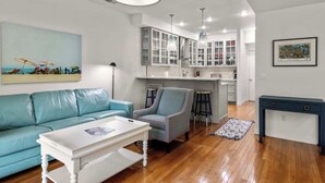 A modern living room with a blue sofa, a white coffee table, and a patterned armchair. There is a kitchen area with white cabinets and a bar stool. - Midtown Riverfront Condo Extended Stays (Traverse City)