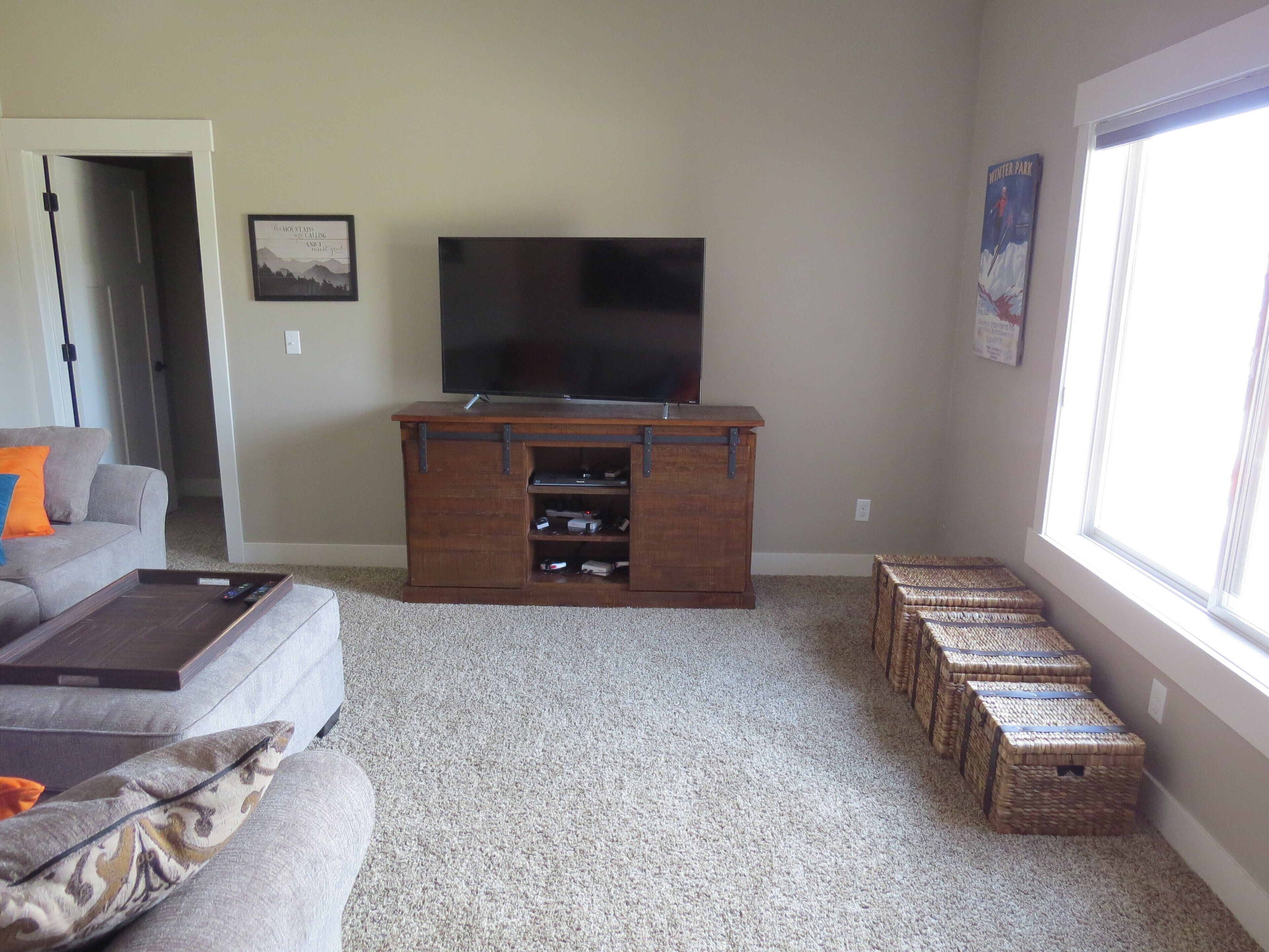 A living room with a flat-screen TV on a wooden stand, a gray sofa with an orange pillow, a coffee table, and wicker baskets against the wall.
