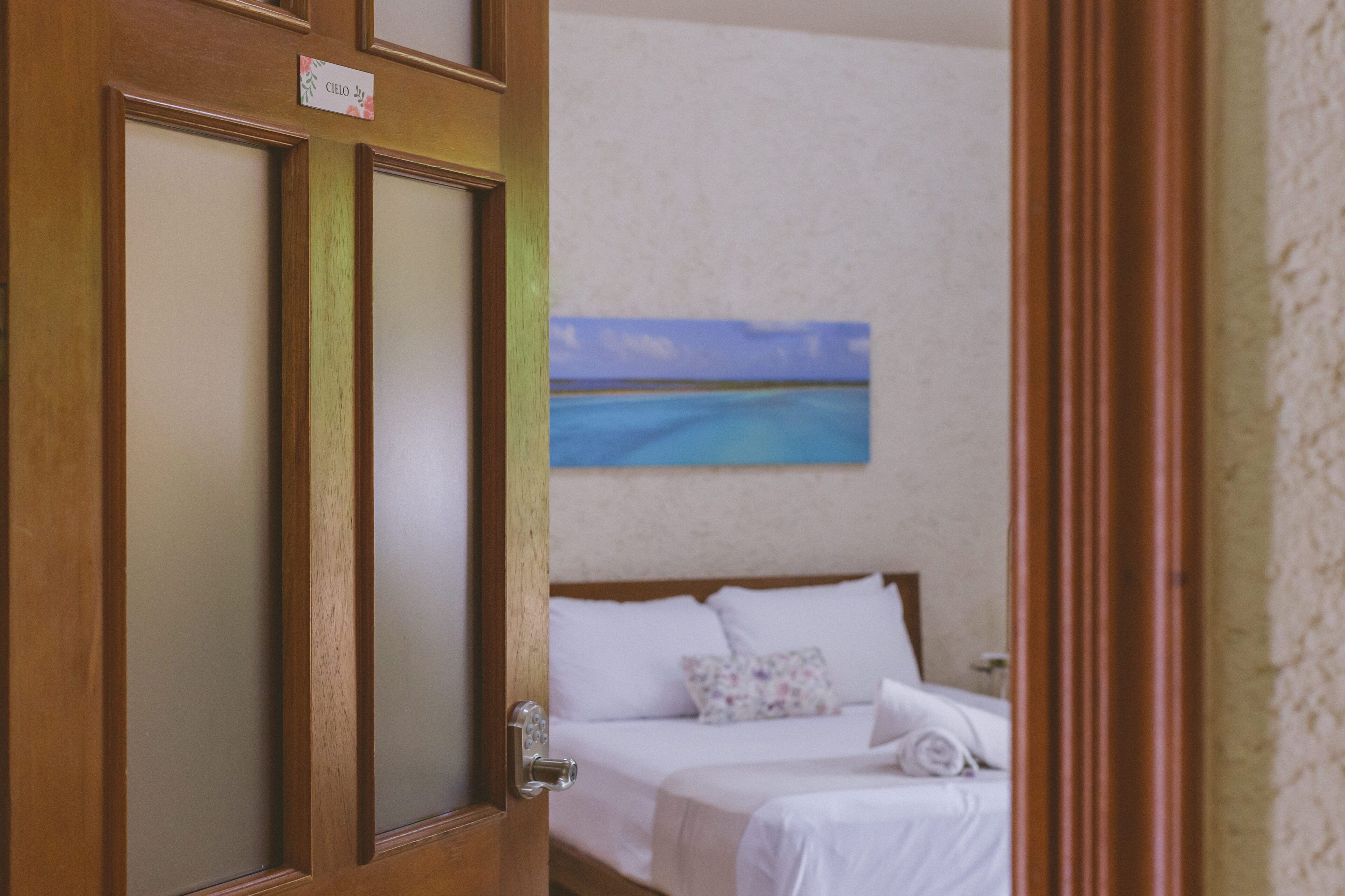 Una camera d'albergo con una porta in legno, un letto con lenzuola bianche, un quadro raffigurante una spiaggia e un interruttore della luce montato a parete.