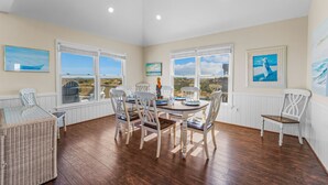 A dining area with a view of the landscape, white chairs, a glass table, and artwork on the walls. - Oceanfront Home w/ Pool, Hot Tub, Game Room, Tiki Bar, Community Pool and Tennis (Waves)