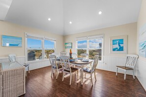Un espace repas offrant une vue sur le paysage, avec des chaises blanches, une table en verre et des œuvres d’art accrochées aux murs.