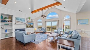 A spacious living room with a high ceiling, wooden beams, and large windows offering a view of the beach. - Oceanfront Home w/ Pool, Hot Tub, Game Room, Tiki Bar, Community Pool and Tennis (Waves)