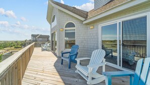 A wooden deck with Adirondack chairs, a glass door leading to a covered area, and a view of the landscape. - Oceanfront Home w/ Pool, Hot Tub, Game Room, Tiki Bar, Community Pool and Tennis (Waves)