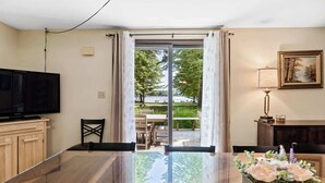 A modern living room with a flat-screen TV, a dining area with a table and chairs, and a view of a garden through a glass door. - Peaceful Lakefront Home with Amazing Outdoor Space (Pullman)