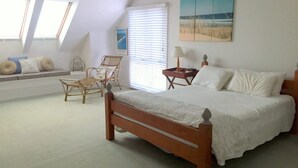 Een slaapkamer met een houten bed, een rieten stoel, een bijzettafeltje met een lamp en een raam met jaloezieën.
