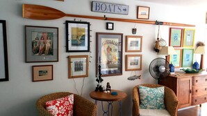 Een kamer met een houten bord aan de muur waarop 'BOATS' staat, verschillende ingelijste foto's, een kleine ronde tafel met een ventilator, rieten stoelen en een houten kast.