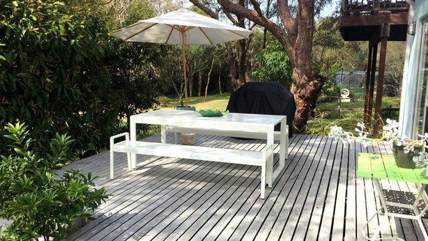 Een terras van hout buiten, met een witte tafel en een bankje, omgeven door groen en een boom.
