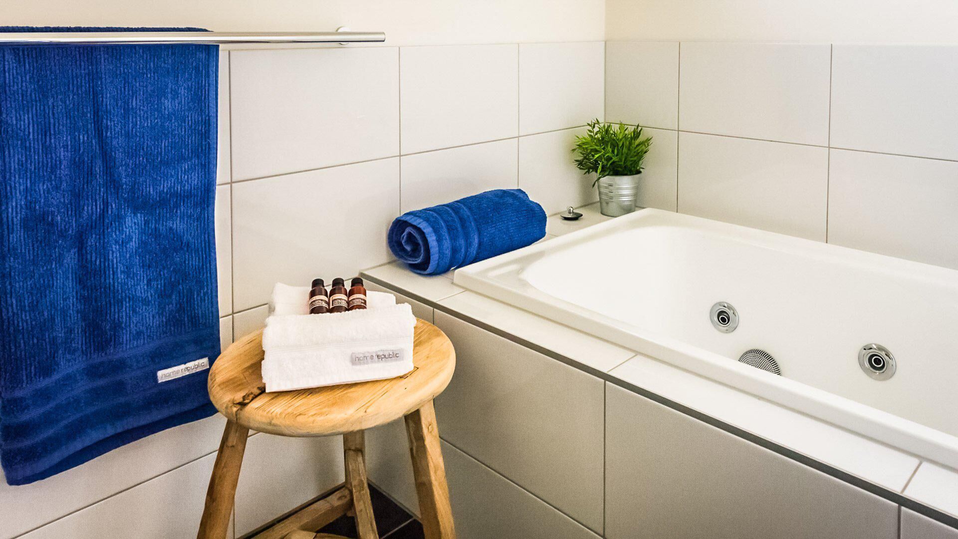Une salle de bain avec une baignoire, un tabouret en bois sur lequel est posée une serviette, une serviette bleue et une plante en pot.