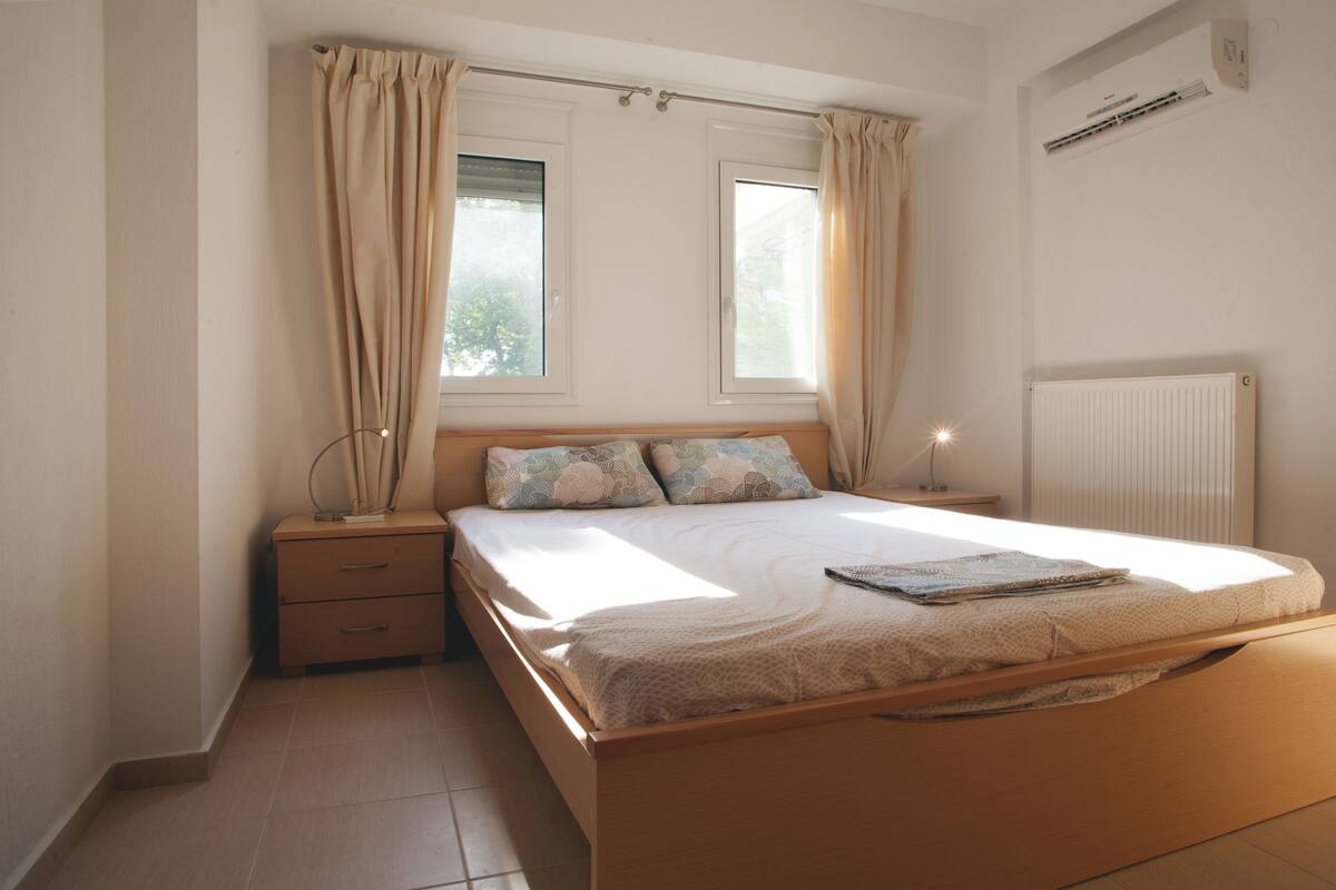 Un dormitorio con una cama de madera, dos ventanas con cortinas, una mesita de noche y un radiador.