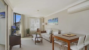 A modern living room with a sofa, coffee table, and TV. There is a large window with curtains, a potted plant, and a painting on the wall.