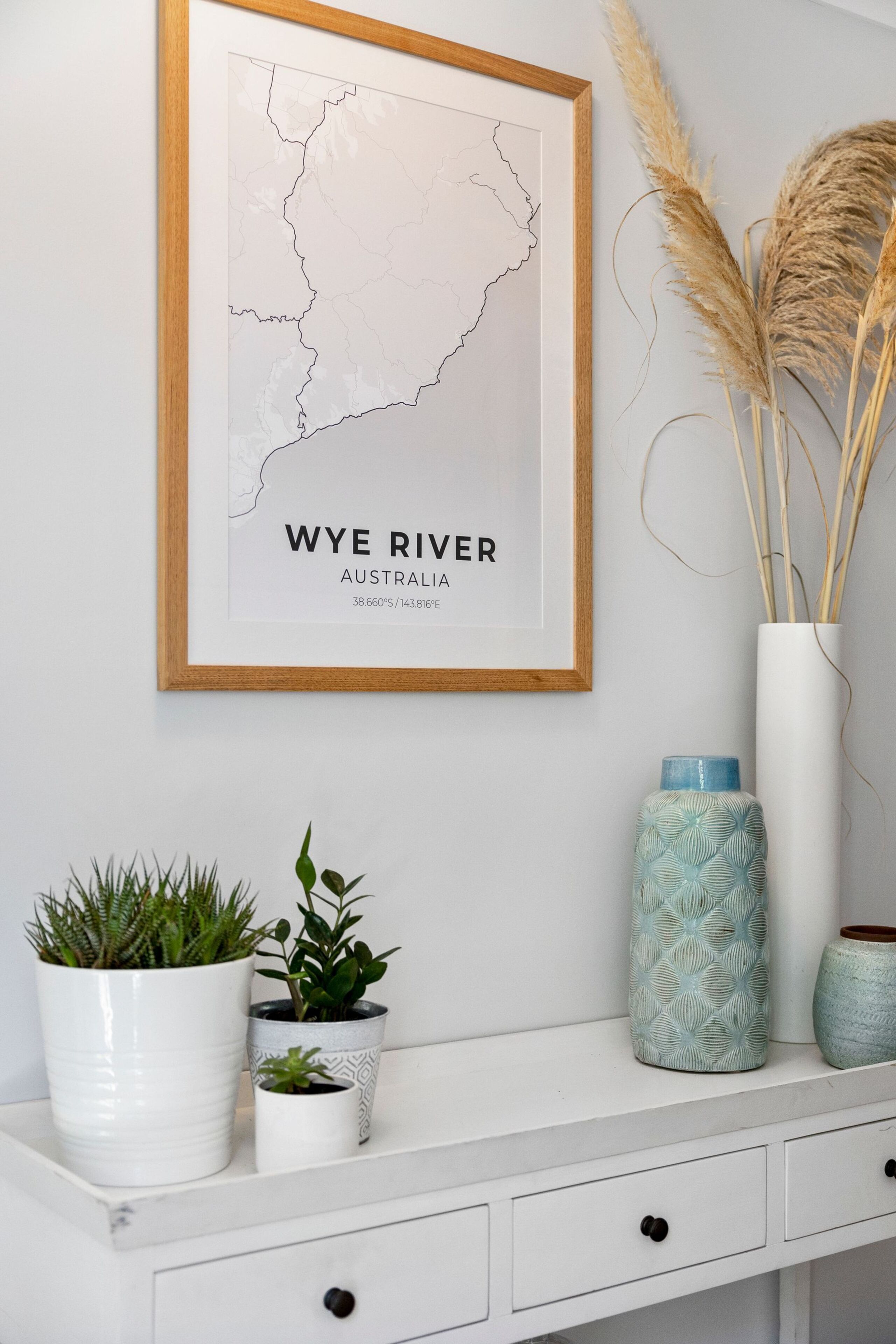 Une carte encadrée de la rivière Wye, en Australie, avec les coordonnées, une console blanche ornée de plantes et d’un vase, et des plumes d’herbe des pampas.