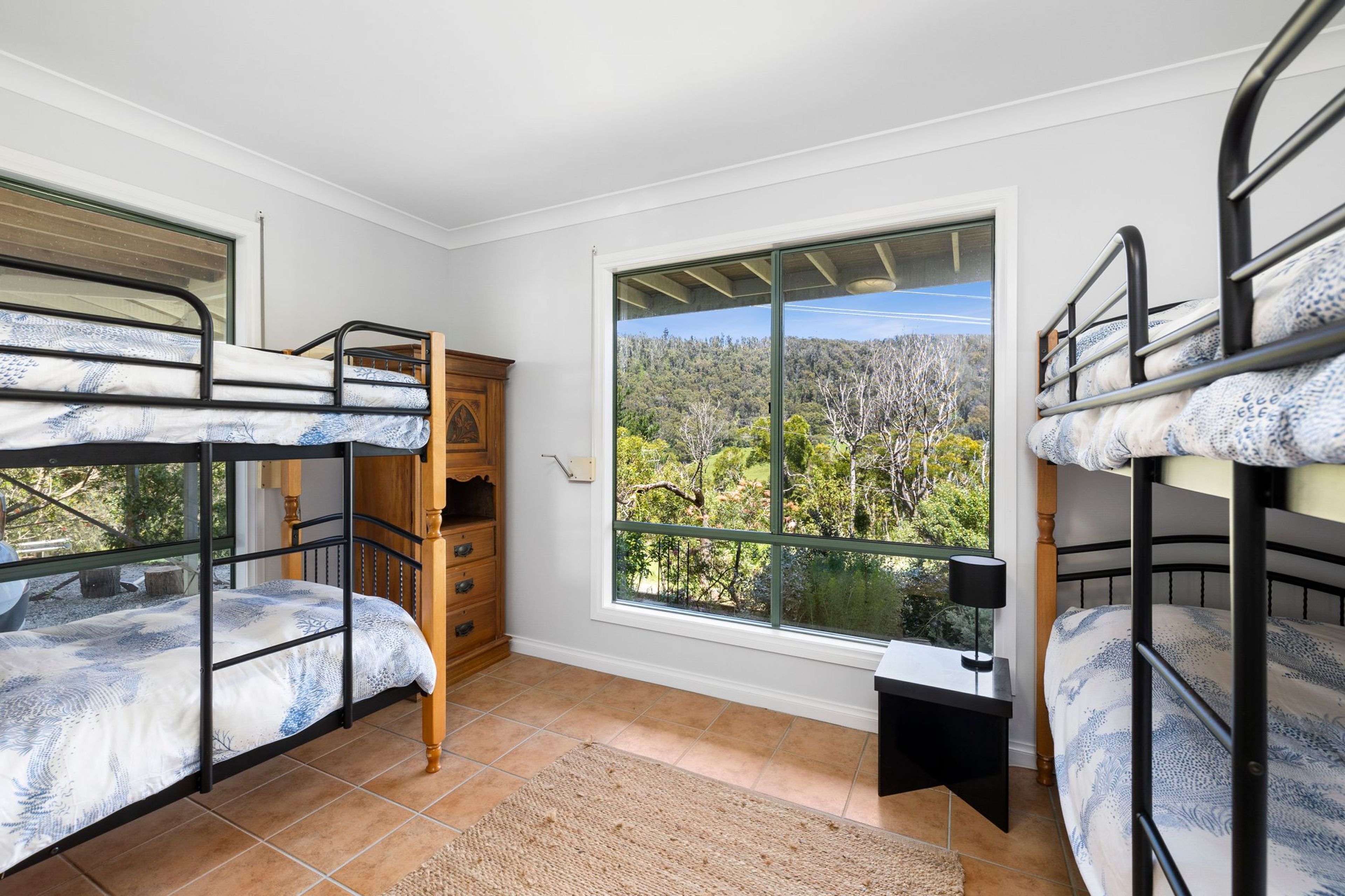 A dormitory room with bunk beds, a wooden dresser, a window with a view of trees, and a tiled floor.
