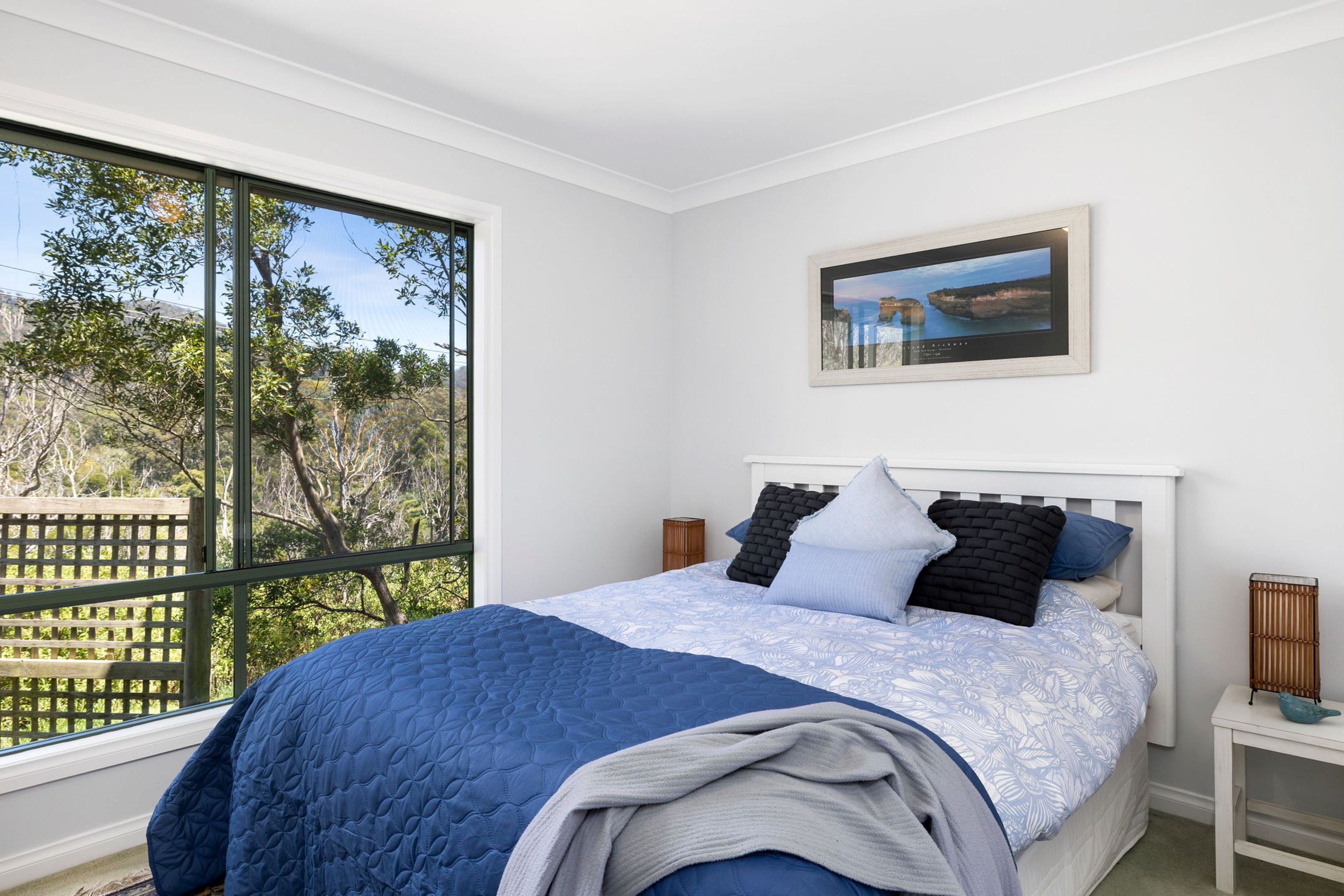 A bedroom with a large window, a bed with a blue quilt, and a framed picture on the wall.