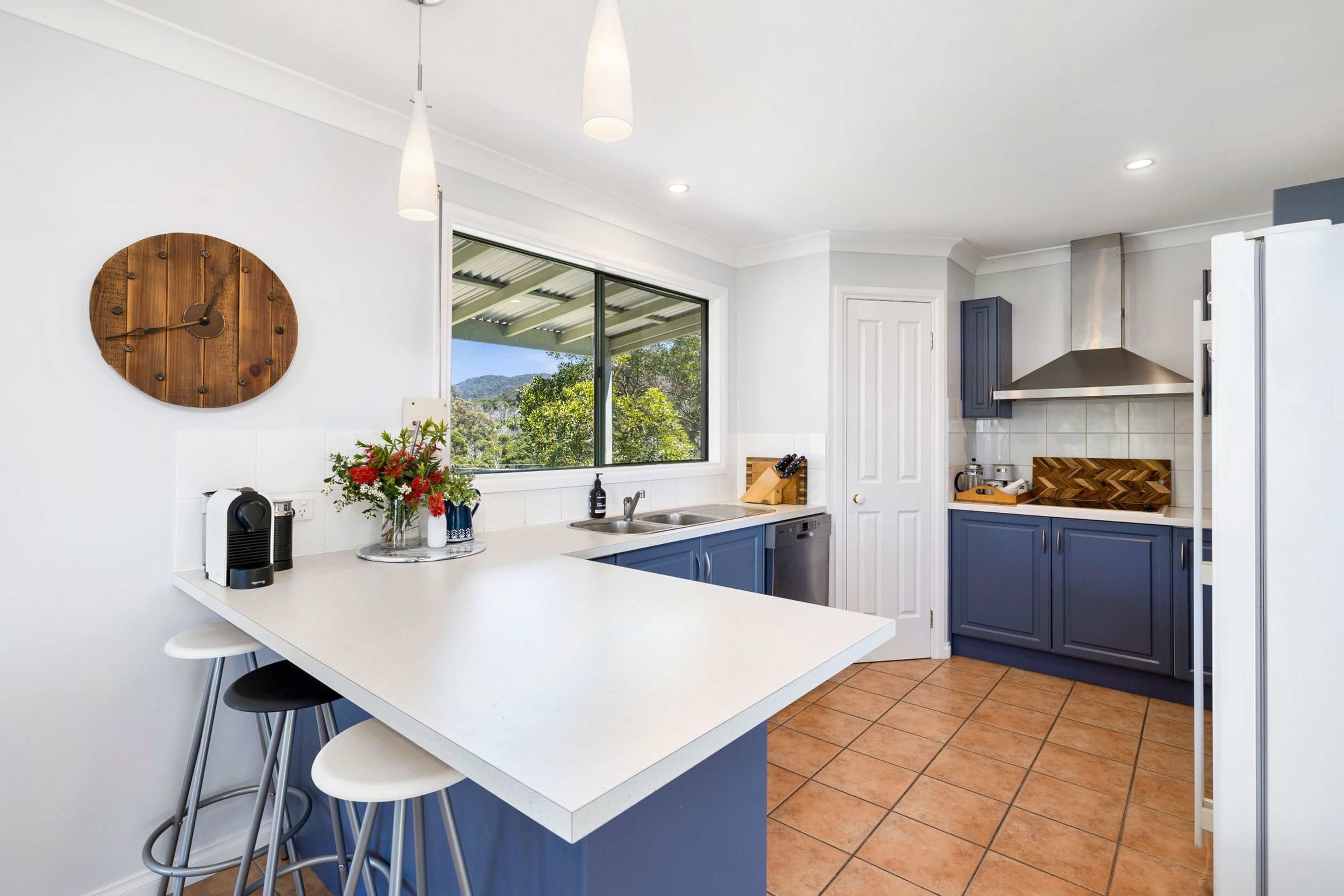 A modern kitchen with a large island, bar stools, a wooden clock, and a view of the outdoors.