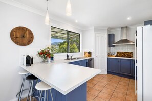 A modern kitchen with a large island, bar stools, a wooden clock, and a view of the outdoors. - Tumbaramba (Wye River)
