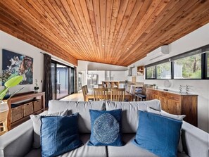 Un soggiorno moderno con soffitto in legno, un divano grigio con cuscini blu, un'area pranzo e una cucina sullo sfondo.