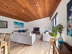 Un soggiorno moderno con soffitto in legno, divani bianchi, televisore e opere d'arte alle pareti.