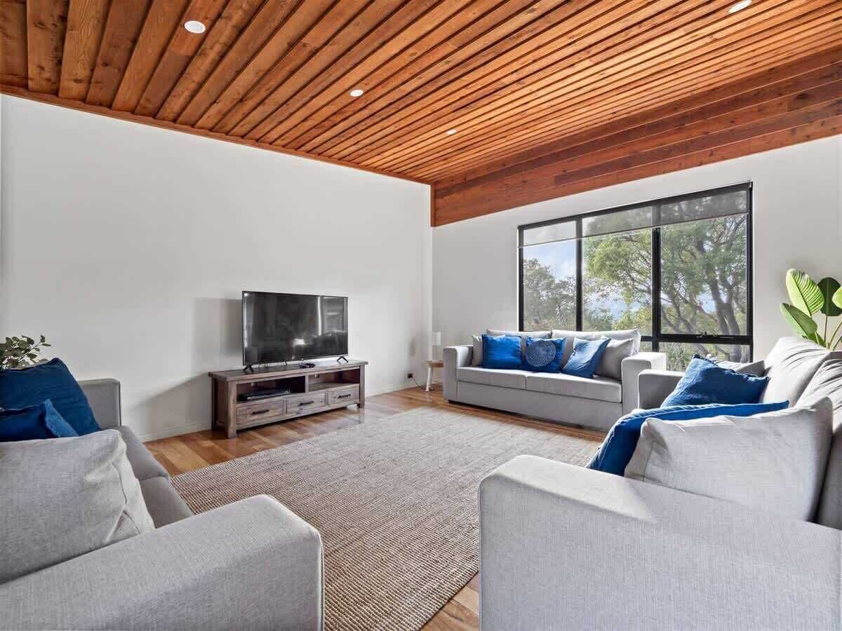 A modern living room with a flat-screen TV, two grey sofas with blue cushions, a wooden ceiling, and large windows.