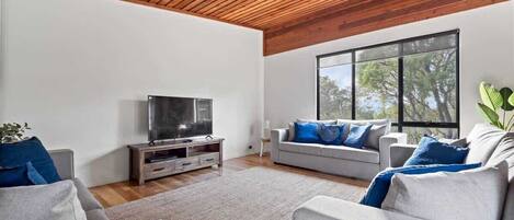 A modern living room with a flat-screen TV, two grey sofas with blue cushions, a wooden ceiling, and large windows.