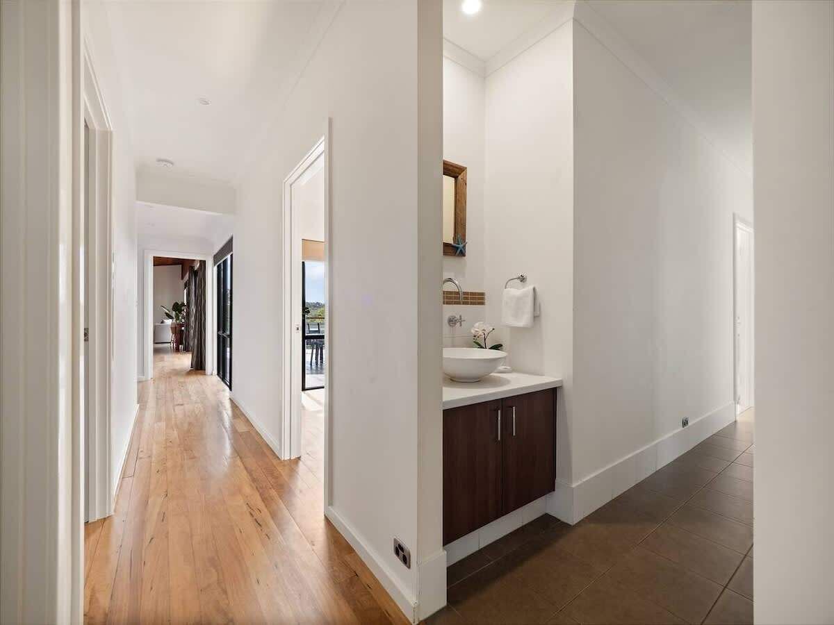 A hallway with wooden flooring, a white wall, and a bathroom with a sink and cabinet.