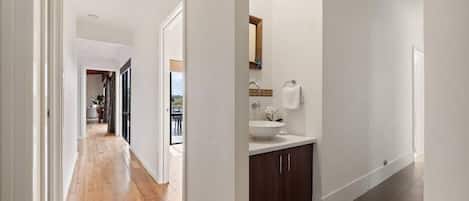 Un pasillo con suelo de madera, pared blanca y un baño con lavamanos y mueble.