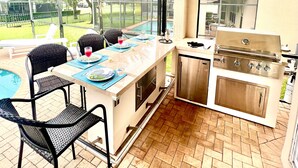 Ein Außenbereich mit Küche, Grill, Esstisch, Stühlen und Blick auf einen Pool.