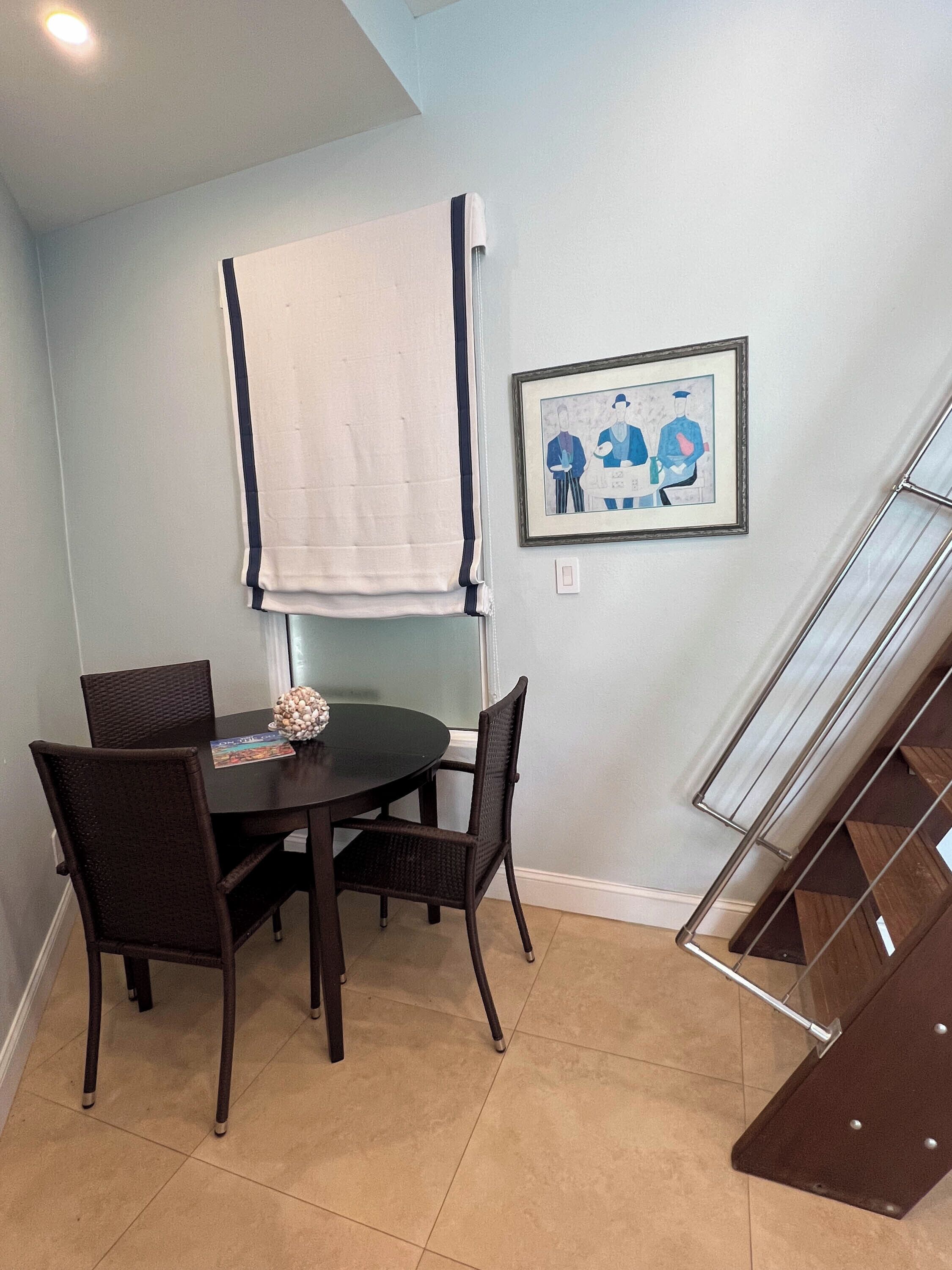 A dining area with a round table, four chairs, a white towel draped over the backrest of the table, and a framed picture on the wall.