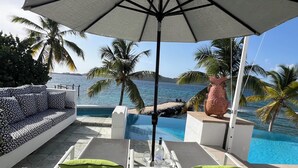 A poolside area with a sofa, two lounge chairs, a table with a bottle of Grey Goose vodka, and a view of the sea and palm trees. - "ONE BEACH ROAD Villa", St Thomas VI, 6 Bedroom, 5 1/2 Bath Beachfront Oasis (St. Thomas)