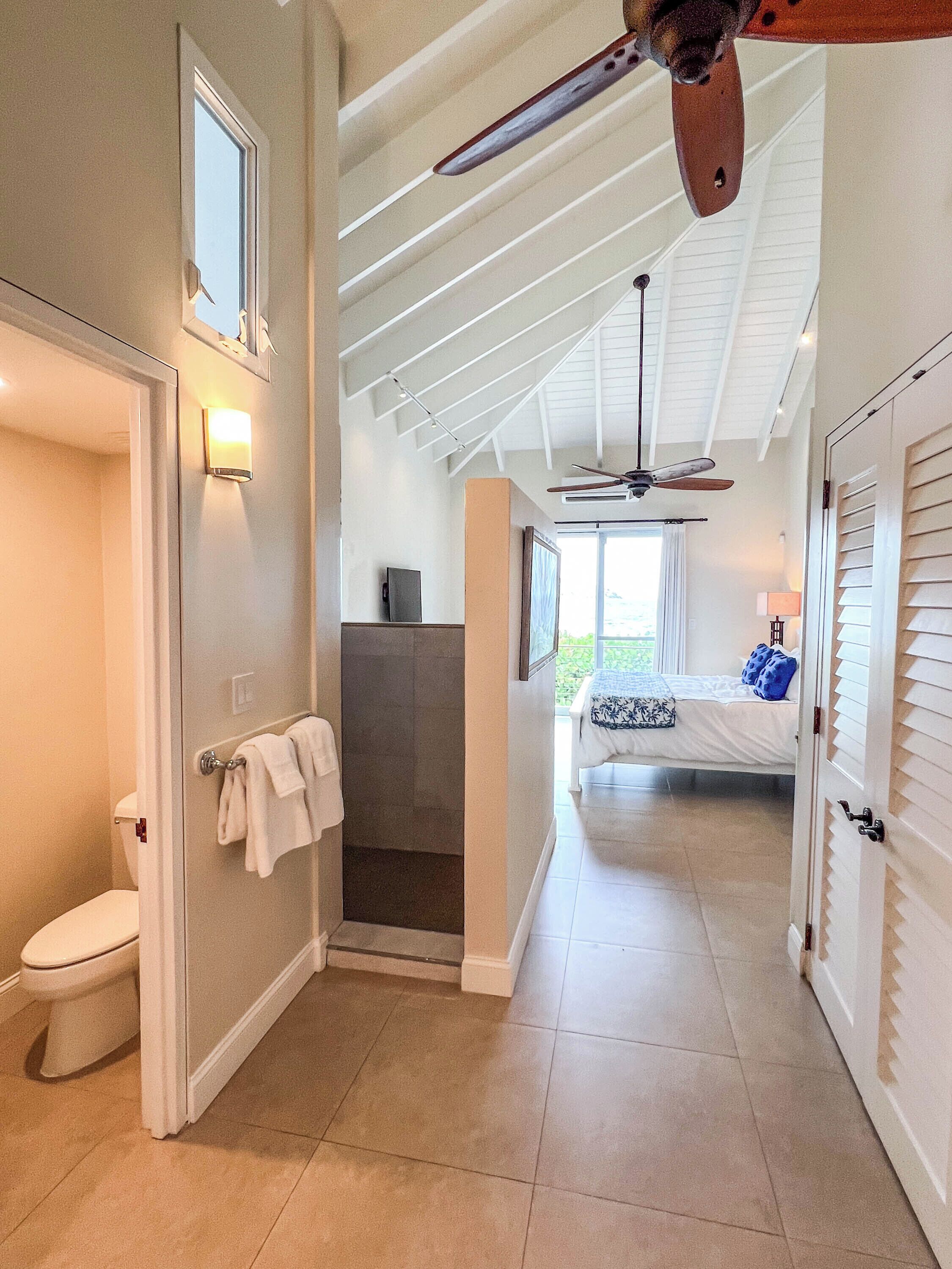 A hallway with a shower, a toilet, a bed with a window, and a ceiling fan.