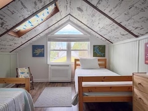 Una camera da letto con soffitto spiovente, una finestra, un letto, una cassettiera e una sedia.