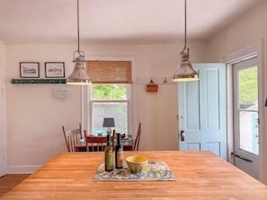 A dining area with a wooden table, two hanging pendant lights, a window with blinds, and a door.