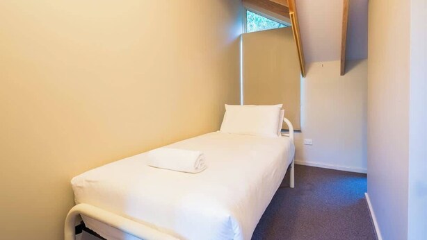 Quarto pequeno com uma cama de solteiro, mesa de cabeceira e janela que oferece luz natural.