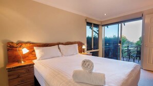 Quarto de hotel com uma cama grande, duas luminárias de cabeceira, cabeceira de madeira, janela com vista e varanda com mobiliário de exterior.