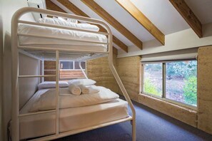 A bunk bed with white bedding, a window with a view of greenery, and exposed wooden beams on the ceiling.