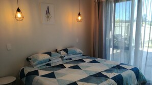 A bed with a geometric patterned bedspread, two wall-mounted pendant lights, a small round table, and a window with sheer curtains. - Family Executive House (Pohara)