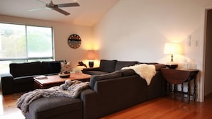 A modern living room with a sectional sofa, a coffee table, and a lamp. There is a ceiling fan, a clock on the wall, and a window with blinds. - Evergreen - spacious family retreat, so close to shops and beaches. (Venus Bay)