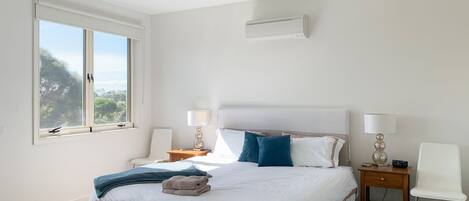 A bedroom with a large bed, two bedside tables with lamps, a window with a view, and a wall-mounted air conditioner.