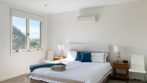 A bedroom with a large bed, two bedside tables with lamps, a window with a view, and a wall-mounted air conditioner.
