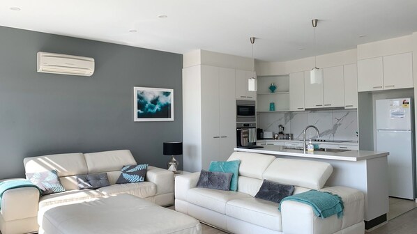 A modern living room with a sofa and ottoman, a kitchen with white cabinets and a marble backsplash, and a grey accent wall with a framed picture.