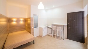 Une pièce de taille modeste comprenant un lit, une petite kitchenette, un bureau avec des chaises et une porte sombre.