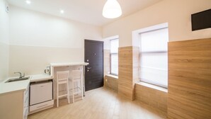 A compact kitchen with a white countertop, a small refrigerator, a sink, and a chair. There is a black door and a window with blinds. - Smart Apartment Zamarstinivska 20a (Lviv)