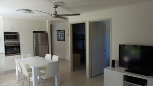 A modern kitchen with white cabinets, a refrigerator, an oven, and a ceiling fan. There is a dining area with a white table and chairs. A television is on a white stand in the corner. - Palmgrove 1 - Rainbow Beach. Gorgeous, modern beach escape in a great location. Aircon. Wifi (RAINBOW BEACH)